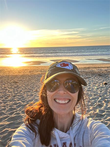 image of woman smiling in Kansas City Chiefs basebase hat and aviator sunglasses on the beach with sunset in background. Long brown hair and grey sweatshirt, taken as a selfie.