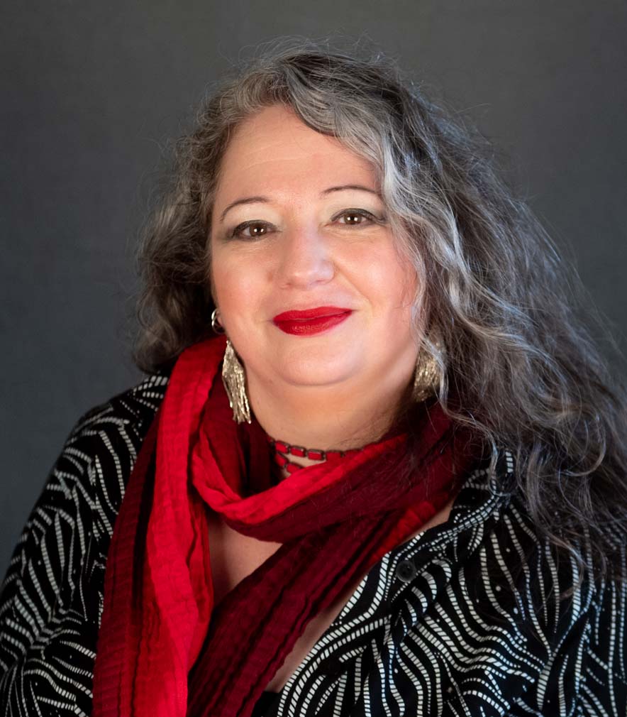 Dr. Sheila Addison A white woman with grey and dark brown hair parted over one eye wearing a black and white printed top and a red scarf. She is smiling at the camera.