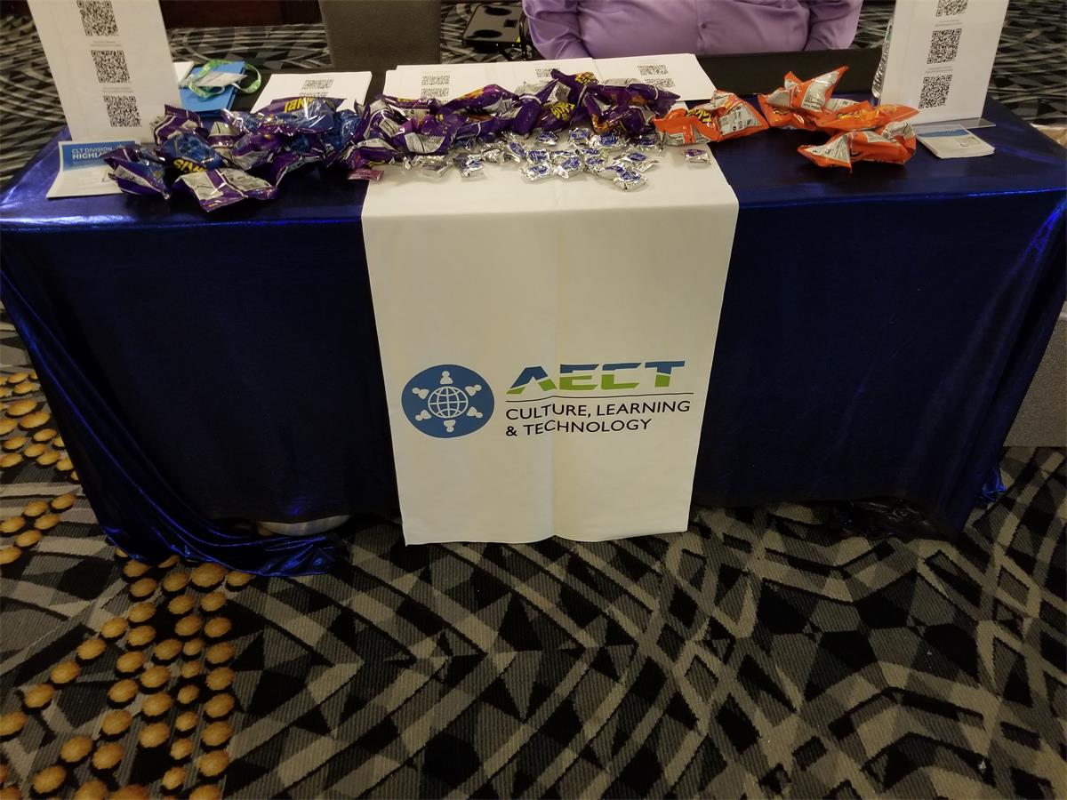 Convention table with AECT drape with satellite looking logo drape and blue lame. On the table are handouts with QR codes, York peppermint patties and Takis