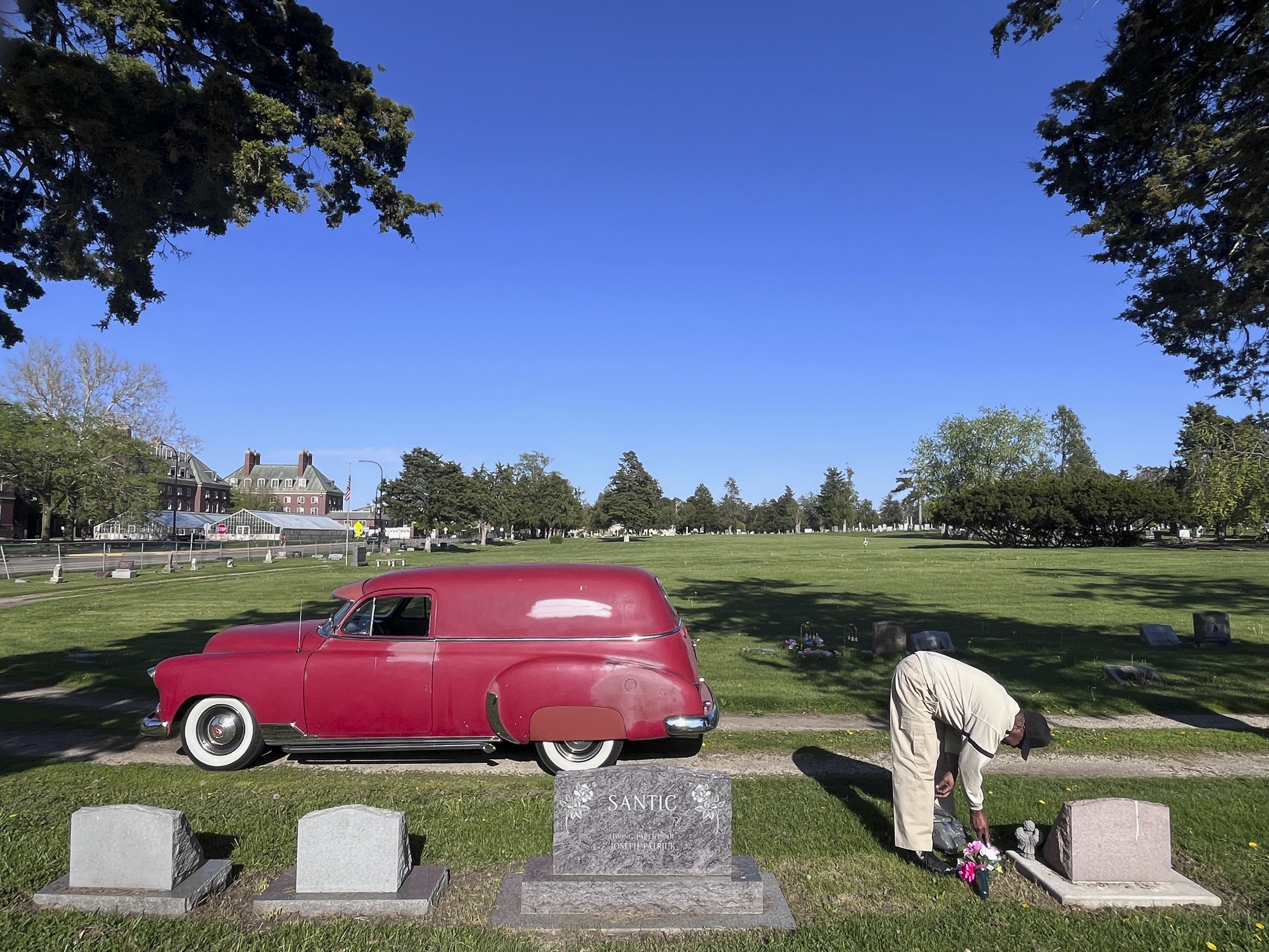 Man placing flowers at cemetery with red car nearby.