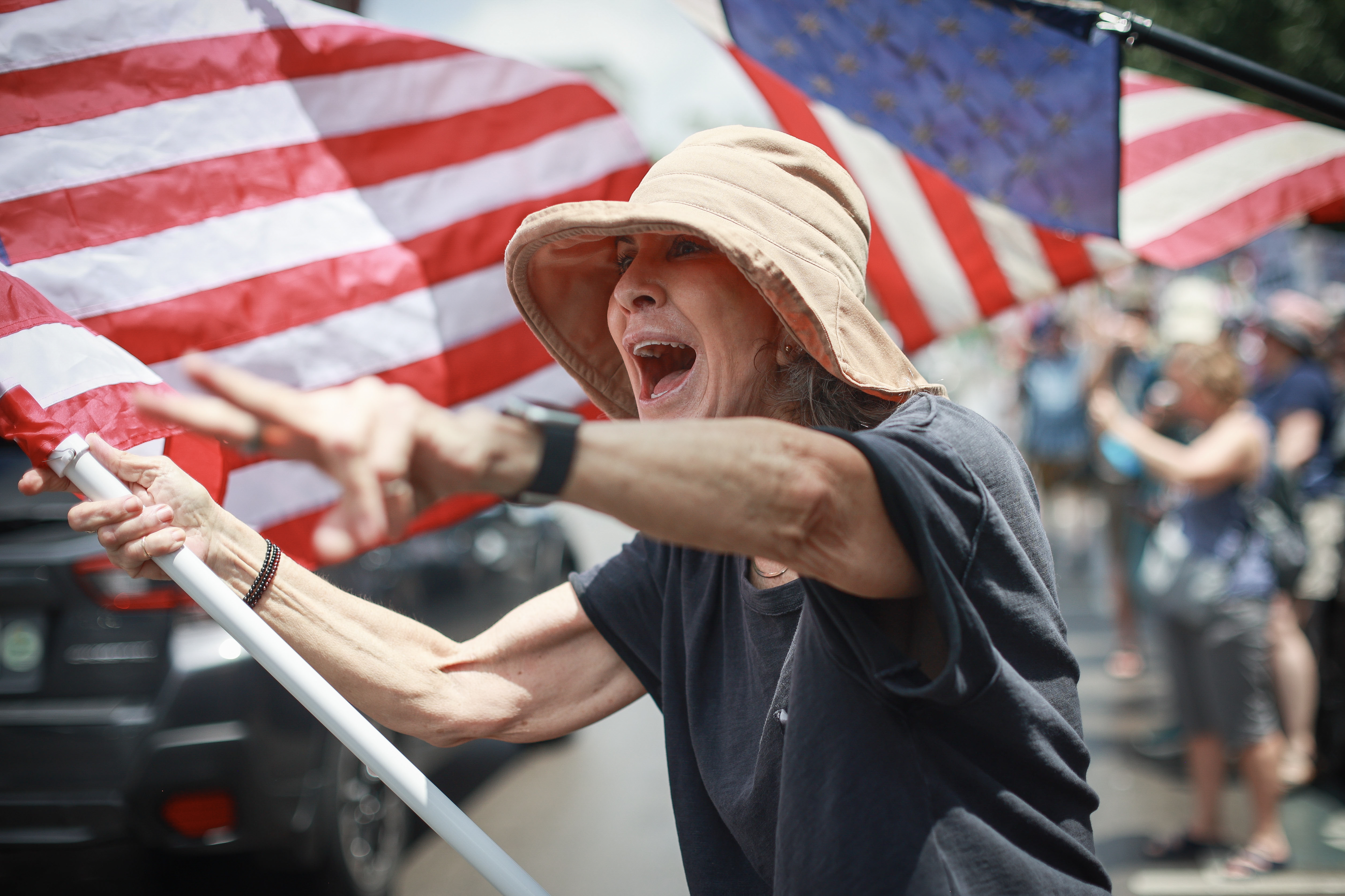 A woman wearing a wide-brimmed hat and holding an American flag yelling and pointing.