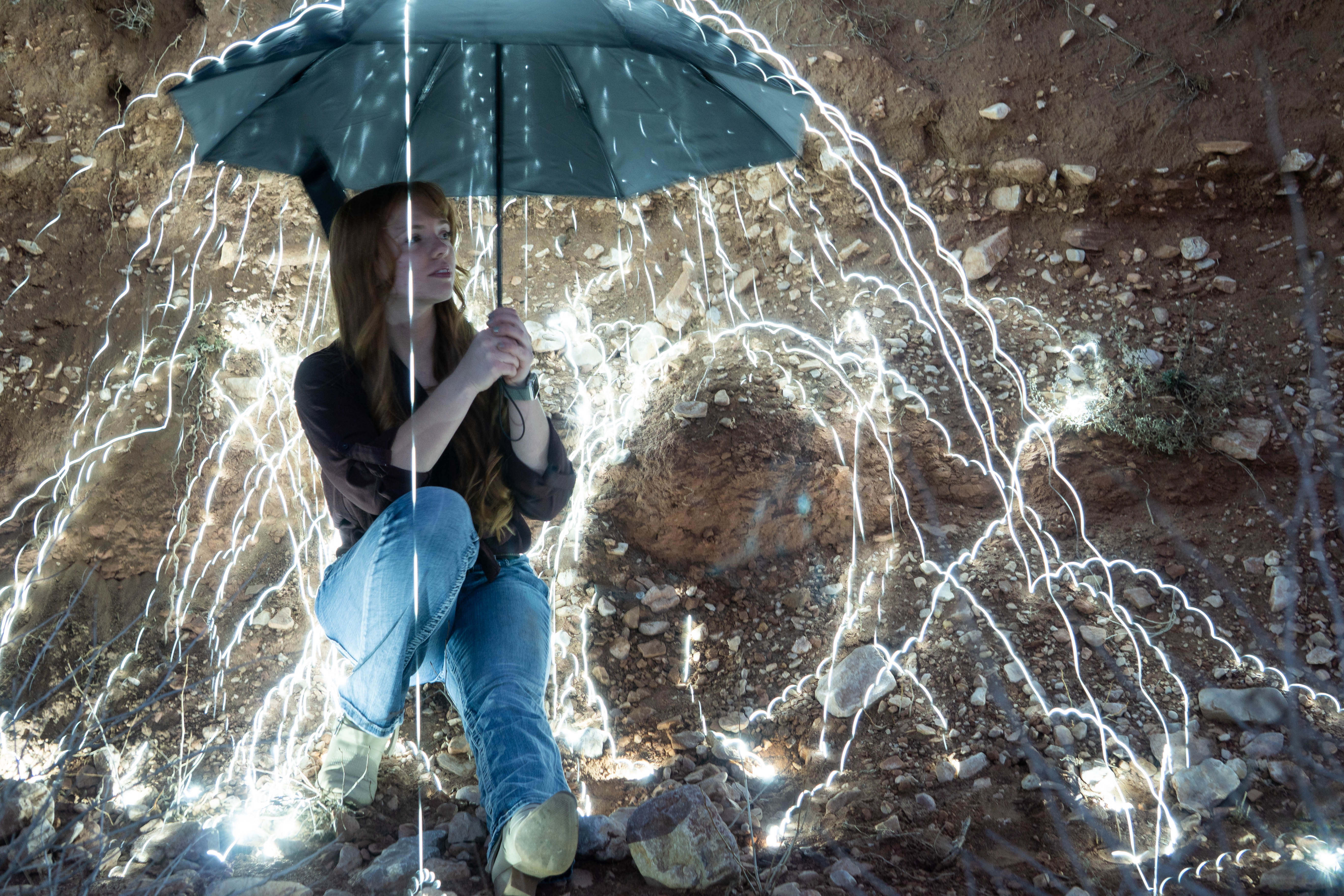 A woman huddles beneath an umbrella against a dirt embankment as trails of light, created by sparklers during a long exposure, cascade around her.