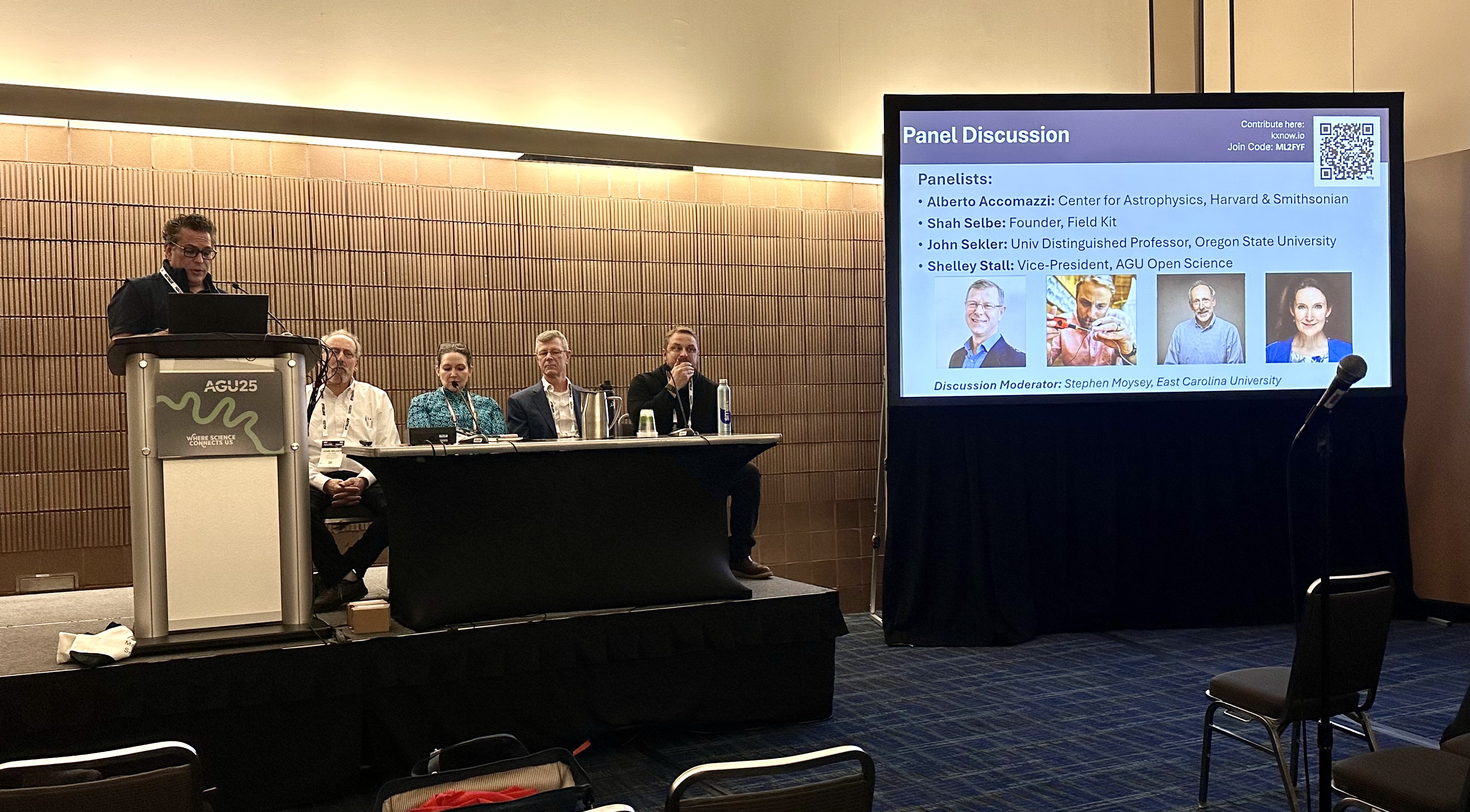 Person standing at a podium on a stage with a panel of people sitting at a table