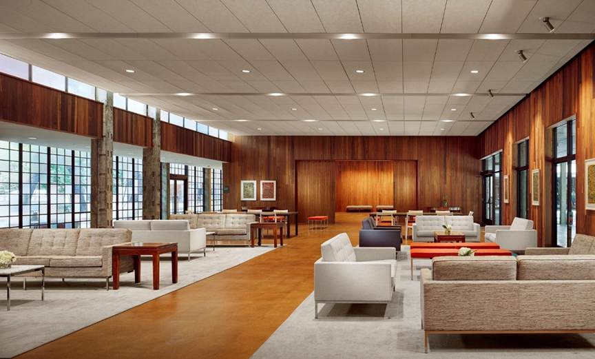 The image depicts a modern, well-lit, and spacious room with a combination of wooden and glass elements, featuring comfortable seating arrangements and a large, open area.

AI-generated content may be incorrect.