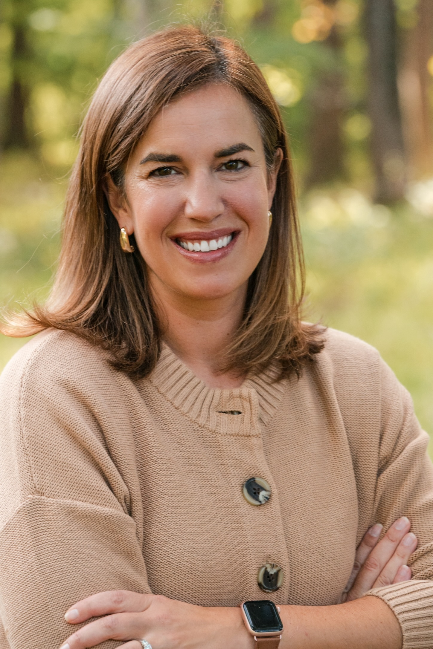The image depicts a smiling brunette woman wearing a beige sweater positioned in a wooded, outdoor setting with trees in the background.