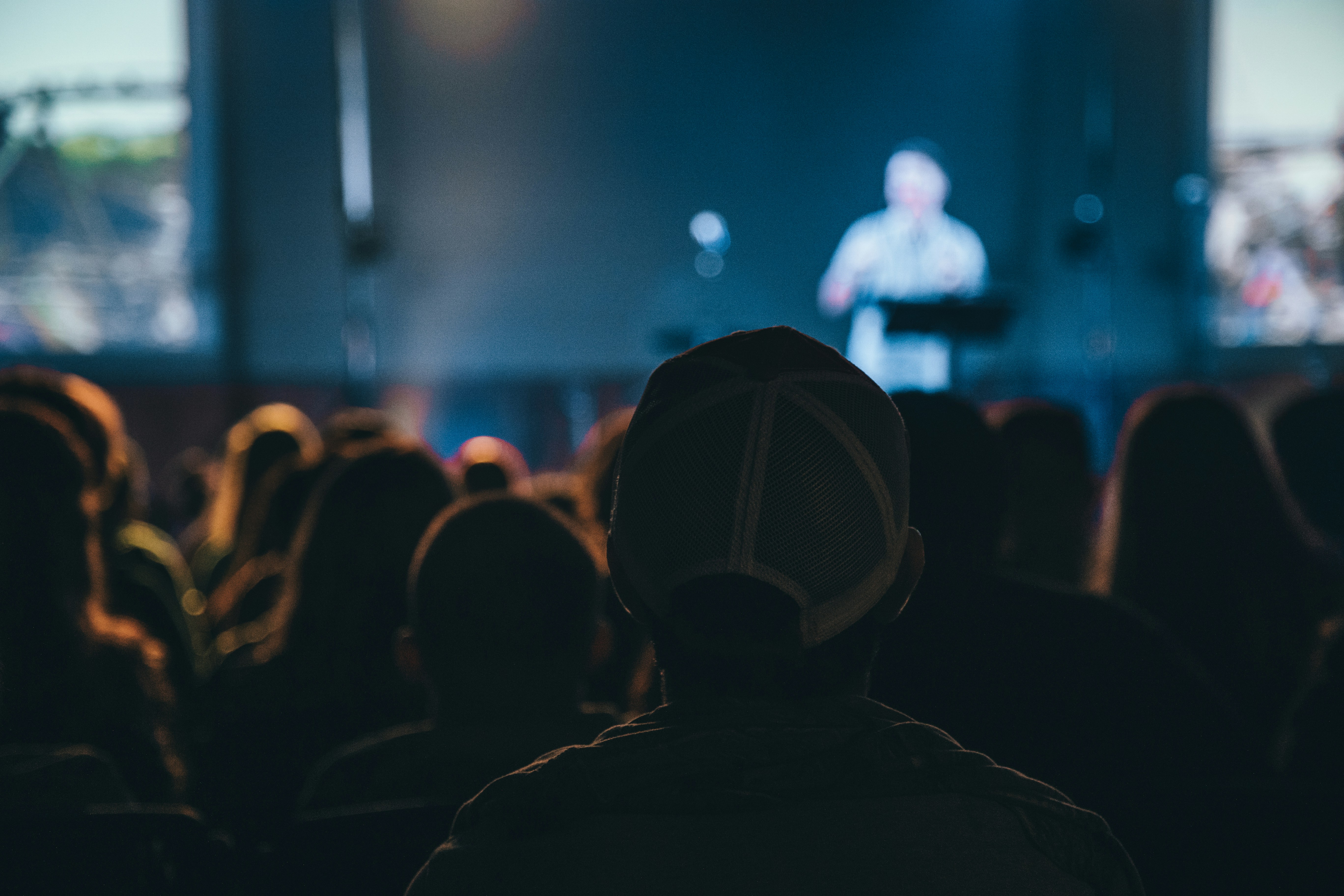 People at a conference in a dark room listening to a presentation. Photo by Terren Hurst on Unsplash Free License
