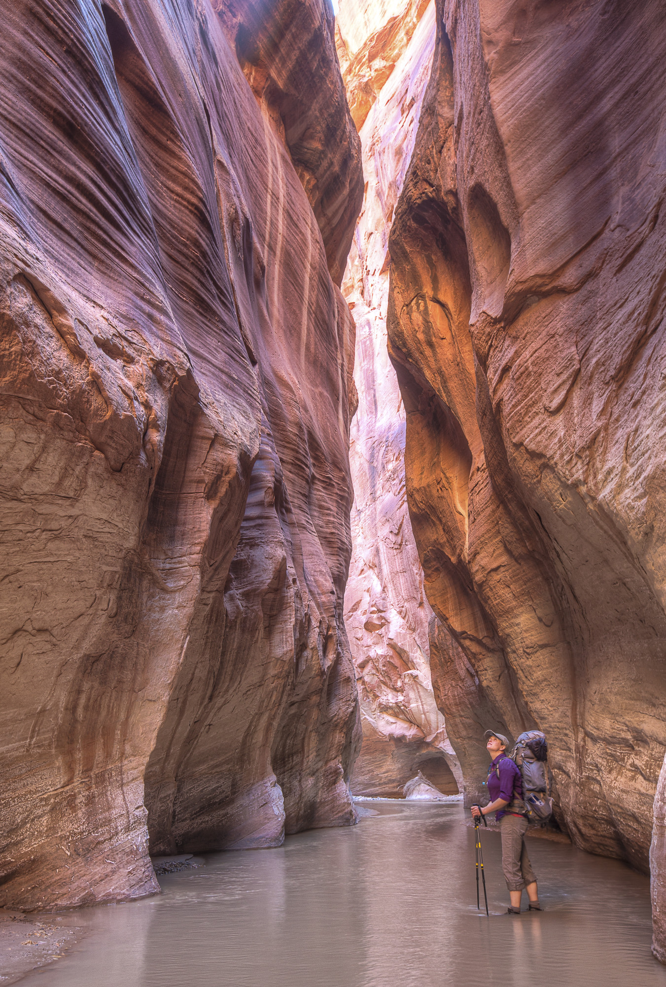 Woman standing at the entrance of an Arizona slot canyon. The early sun casts a shadow at the entrance that makes the rocks appear purple. 