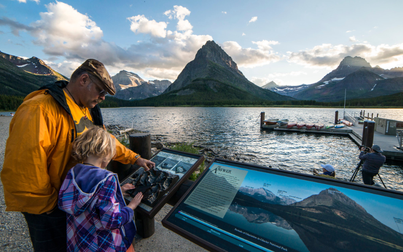 A grandfather and granddaughter read an interpretive sign on the edge of a lake that overlooks the mountains in Glacier National Park 
