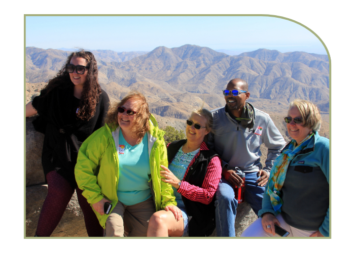 Three women and one man pose for a photo during a sunny day on the summit of a mountain. They are seated with brown mountains behind them. 