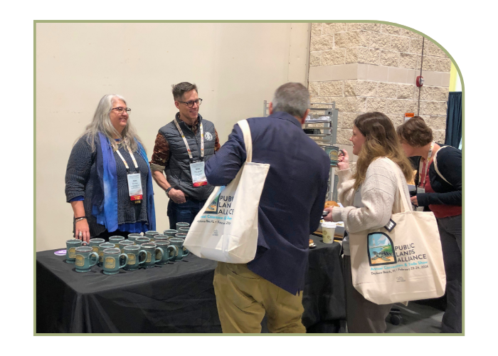 A man and woman are standing behind a table topped with ceramic mugs. They happily chat with a woman who is holding one of the mugs. That same woman is carrying a convnetion tote bag