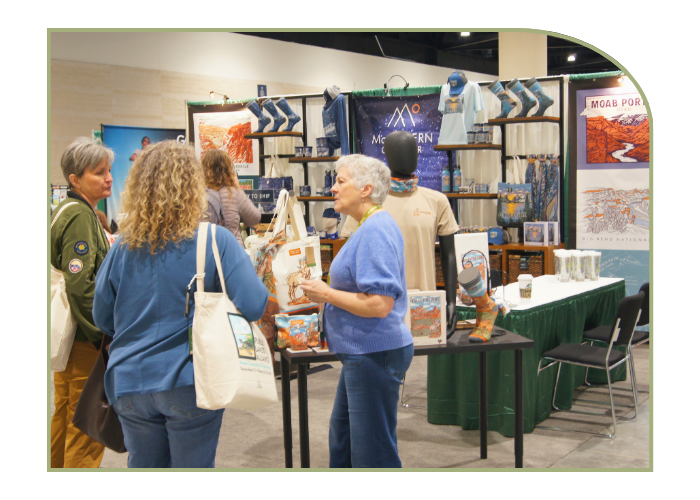 Three women engage in conversation in front of the McGovern Outdoor vendor booth that is stocked with tshirts, bags, socks, and other items related to national parks