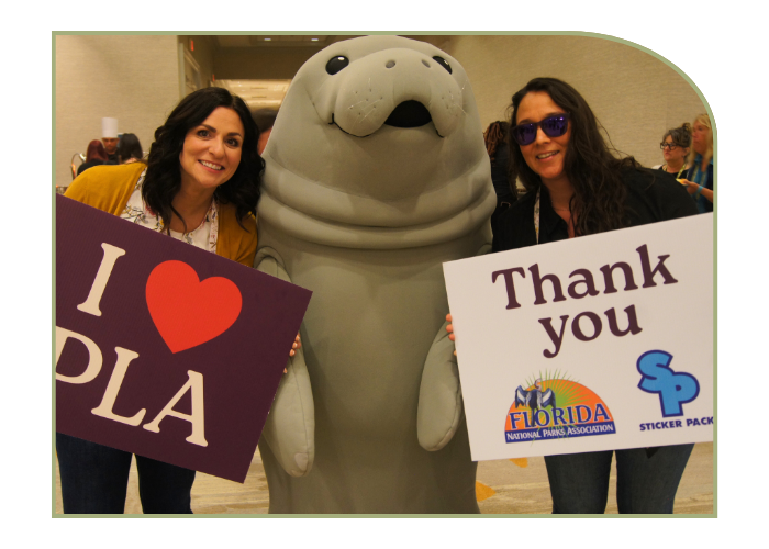two women stand on either side of a very cute manatee costume. They are holding signs and smiling at the camera. One of the signs says "I Heart PLA" the other sign says "Thank you Florida National Park Association and Sticker Pack"