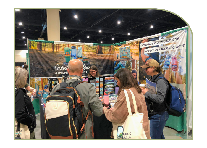 Four people with their backs to the camera are standing at a vendor booth in the trade show. The female vendor has dark hair and glasses. Her water bottles and stickers are nature themed 