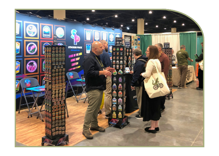 a man and women are in conversation amongst a vendor booth displaying hundreds of nature themed stickers