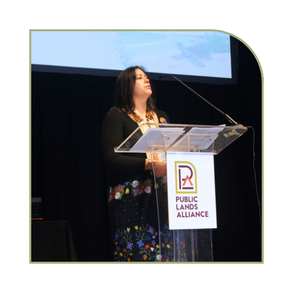a young, dark-haired woman of native american decent stands at a podium with black drapery behind her. The front of the podium has the Public Lands Alliance Logo