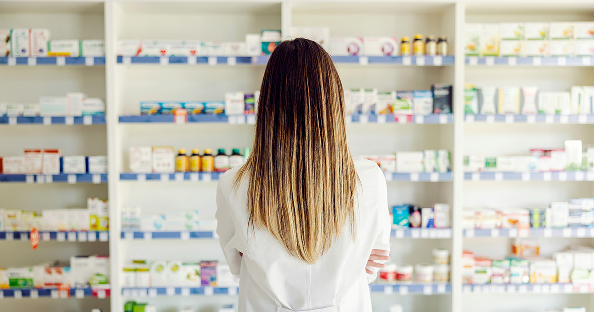 Pharmacist in a white coat viewed from behind, facing shelves stocked with medications in a pharmacy.