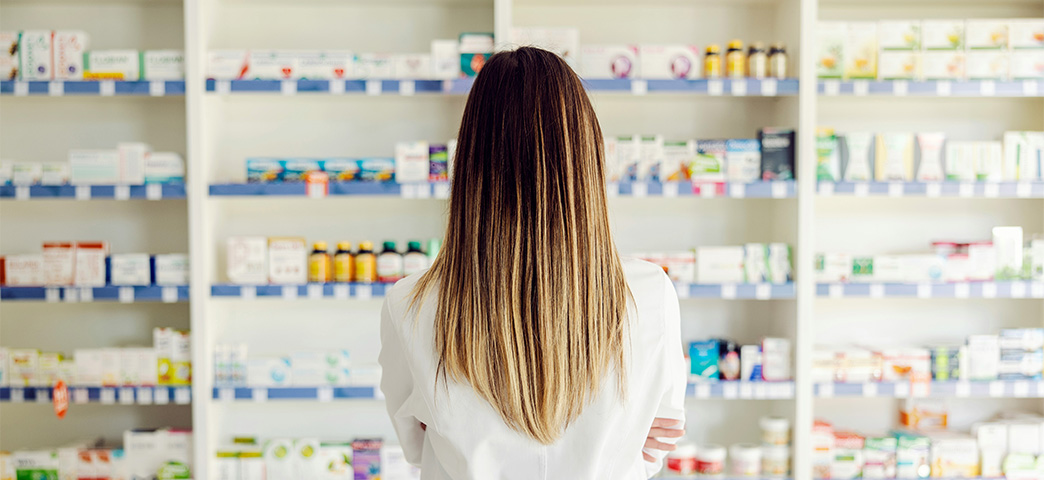 Pharmacist in a white coat viewed from behind, facing shelves stocked with medications in a pharmacy.