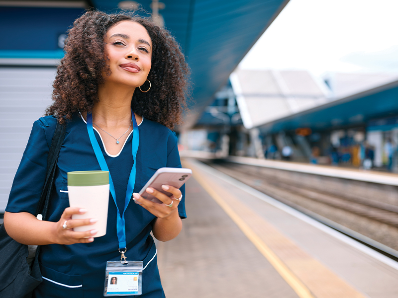 A woman wearing a lanyard holding a coffee and her phone is waiting for a train.