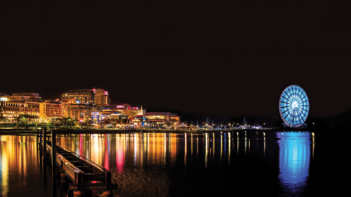 A nighttime panoramic view of National Harbor, Maryland, with colorful city lights reflecting on the Potomac River and a large illuminated Ferris wheel glowing blue on the right.