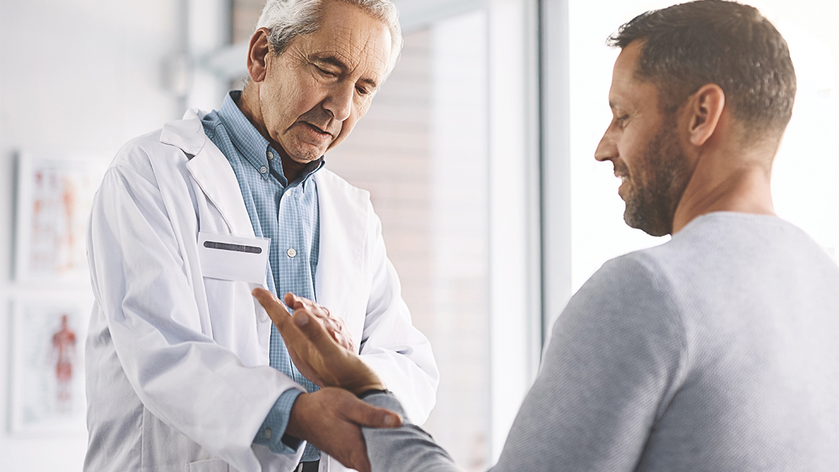 A physician in a white lab coat examines a patient's hand and wrist during a clinical consultation, with anatomical charts visible on the wall in the background.