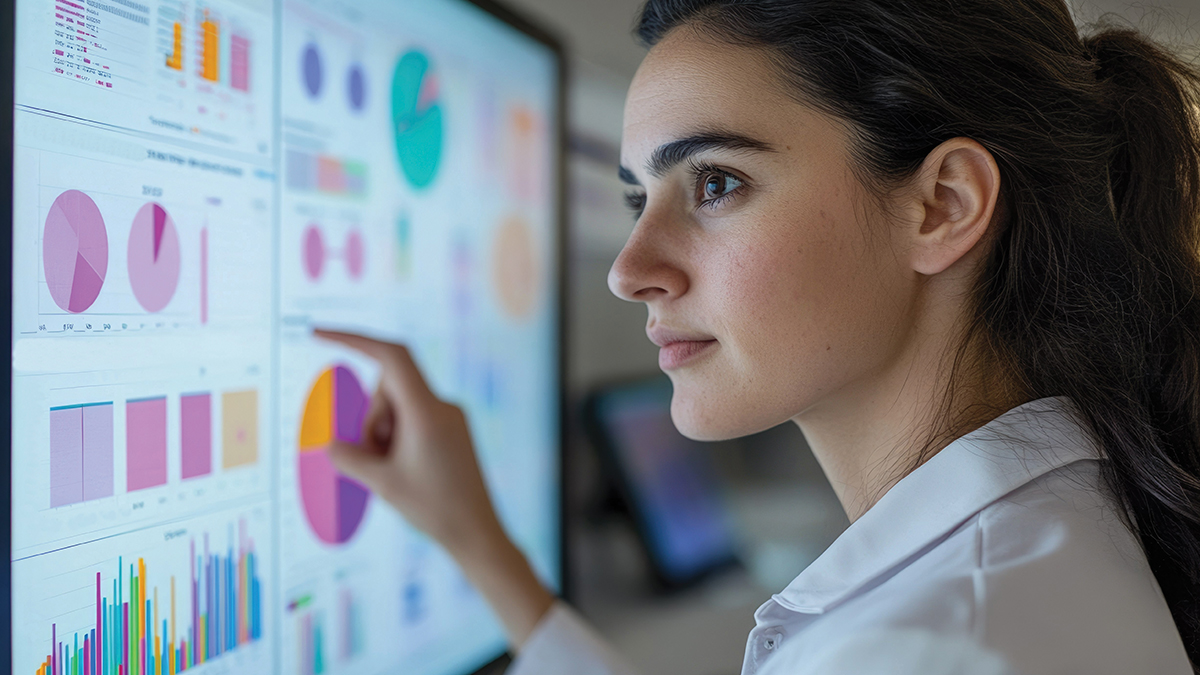 A healthcare professional in a white coat points to a large data dashboard displaying colorful pie charts and bar graphs on a monitor, reviewing performance metrics and analytics.