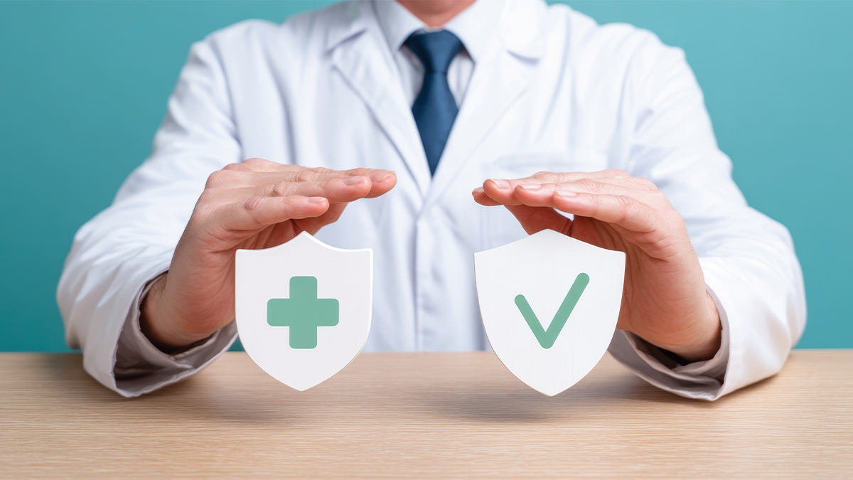 A physician in a white lab coat and blue tie holds his hands over two shield icons on a desk, one displaying a green cross and the other a green checkmark, representing healthcare protection and quality assurance.