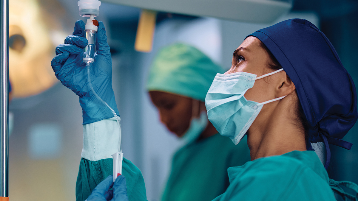 A masked healthcare provider in teal scrubs and blue gloves adjusts an IV drip in an operating room, with a second masked provider visible in the background.
