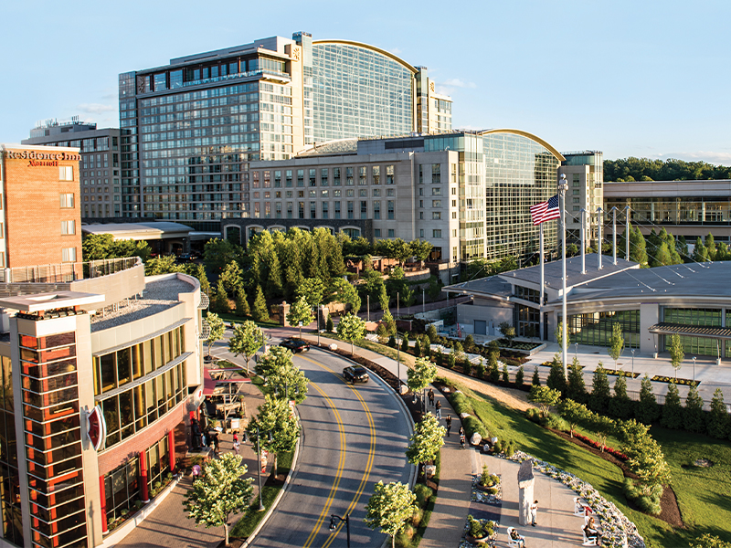 An aerial view of the Gaylord National Resort & Convention Center at National Harbor, Maryland, with its distinctive curved glass roof, an American flag, surrounding hotels, tree-lined streets and landscaped grounds.