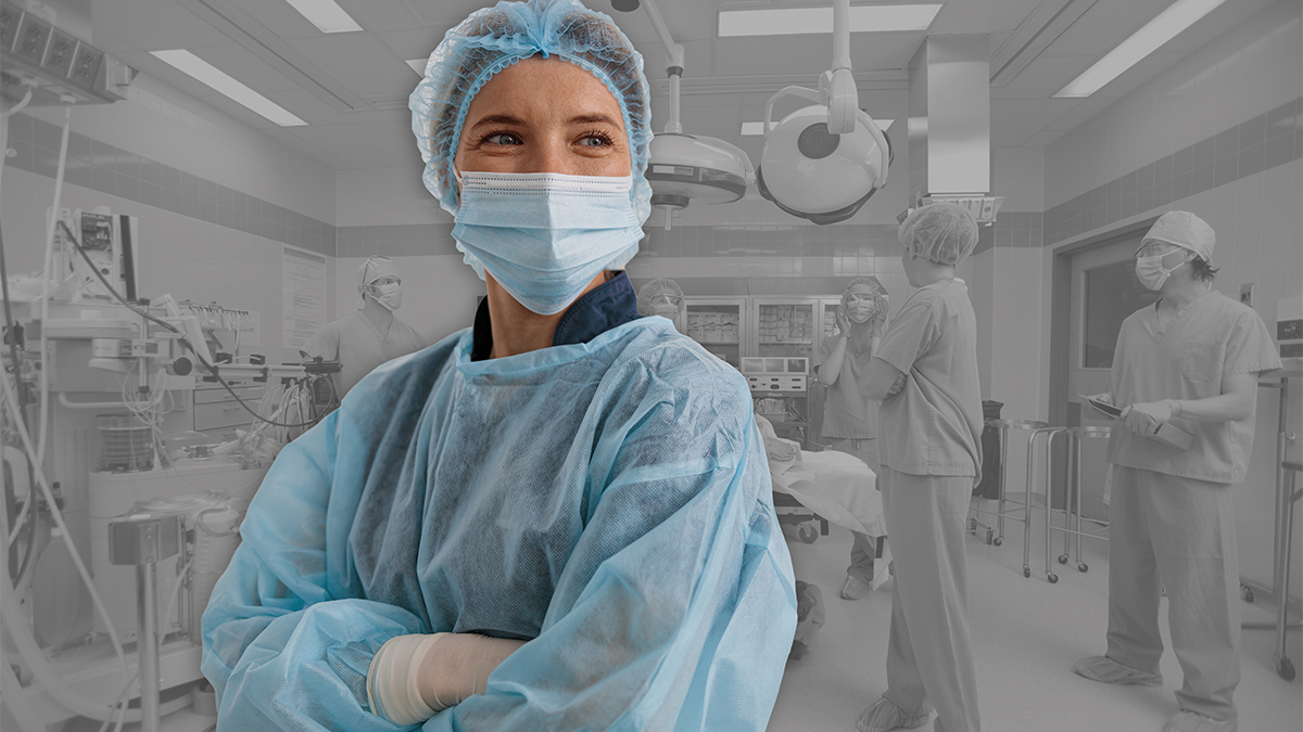 A confident healthcare professional in surgical scrubs, a blue gown, mask, hair net and gloves stands with arms crossed in the foreground, while a surgical team works in a softly faded operating room in the background.
