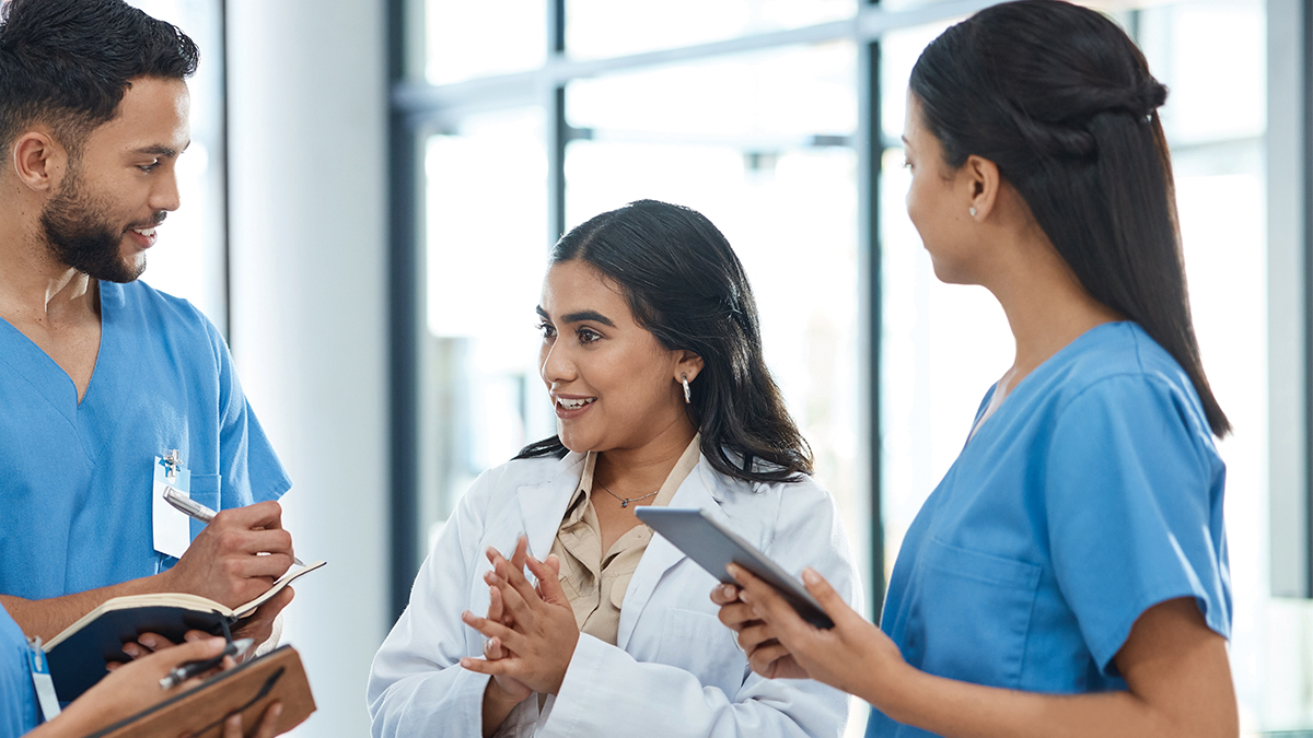 Three healthcare professionals engage in discussion, with one holding a tablet in a bright clinical hallway.