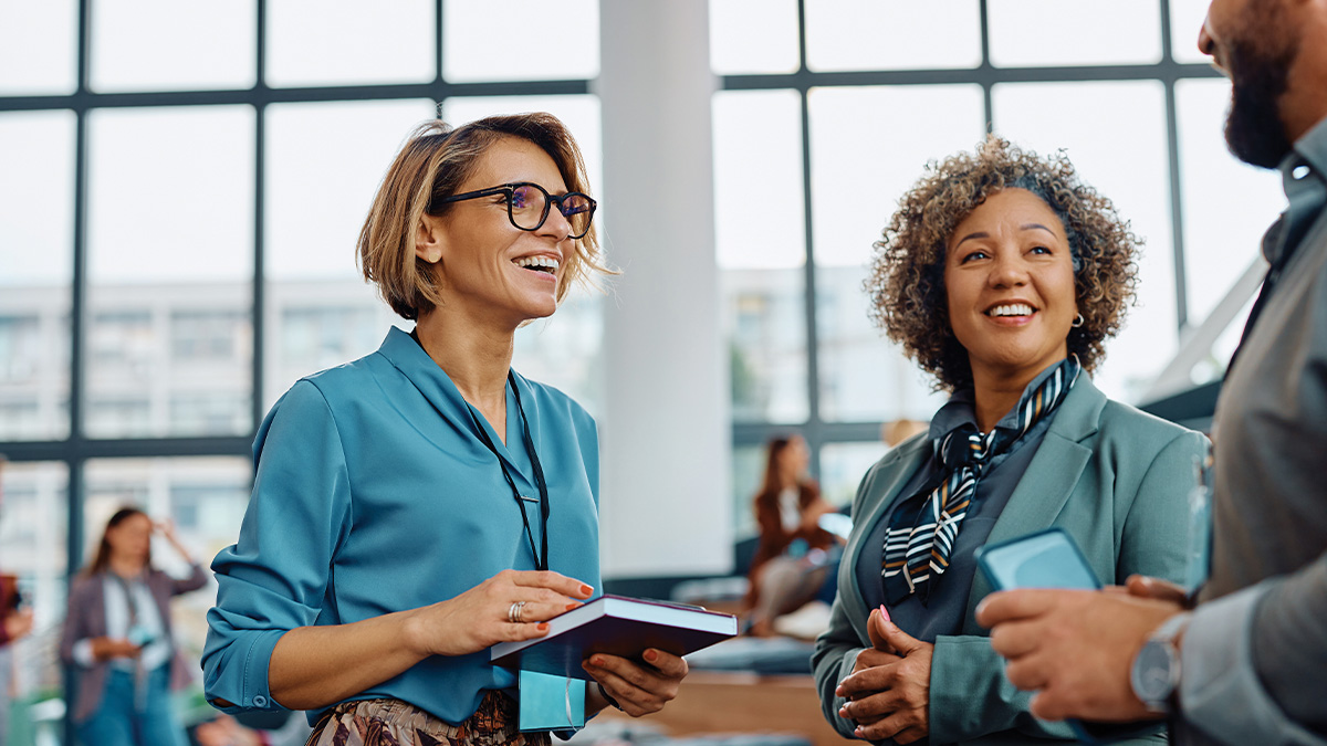Two women wearing conference lanyards smile and chat with a colleague in a bright conference setting.Two women wearing conference lanyards smile and chat with a colleague in a bright conference setting.