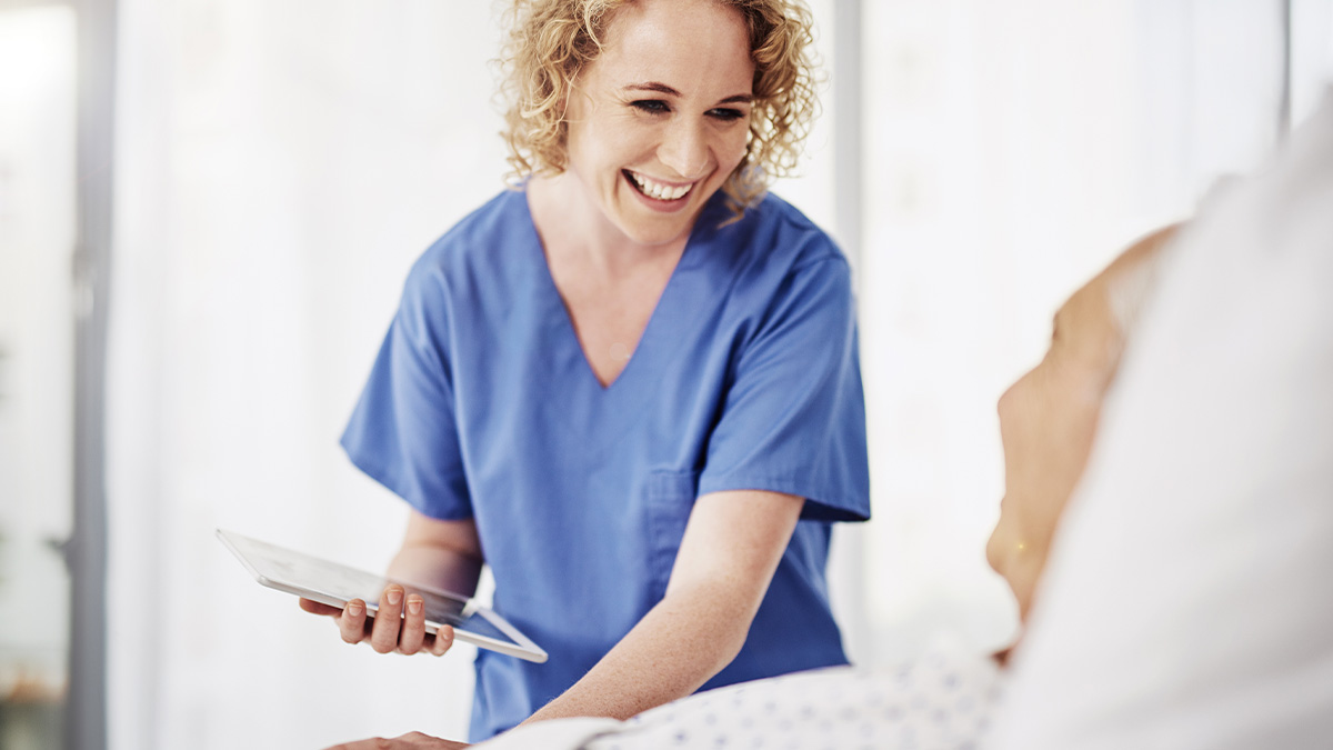 A smiling nurse in blue scrubs reviews information on a tablet while attending to a patient.