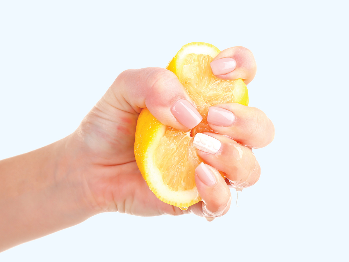 A hand squeezing a lemon half, with juice dripping from the fruit, on a light gray background.