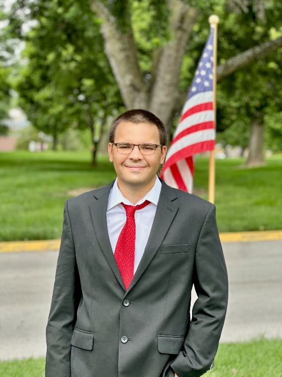 A person in a suit and tie standing in front of a flag

Description automatically generated
