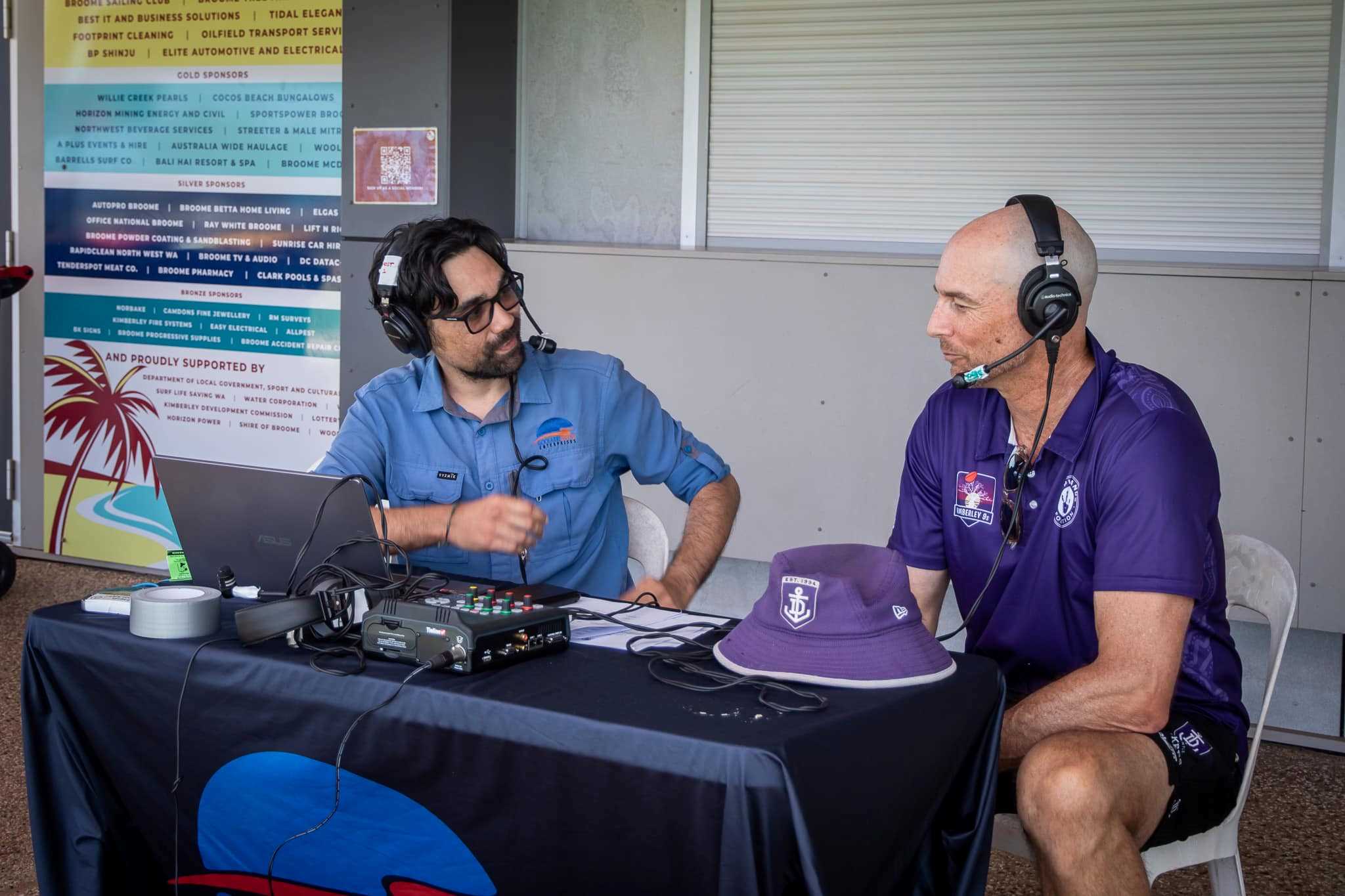 Two people sitting behind a desk with radio controls at an outside broadcast