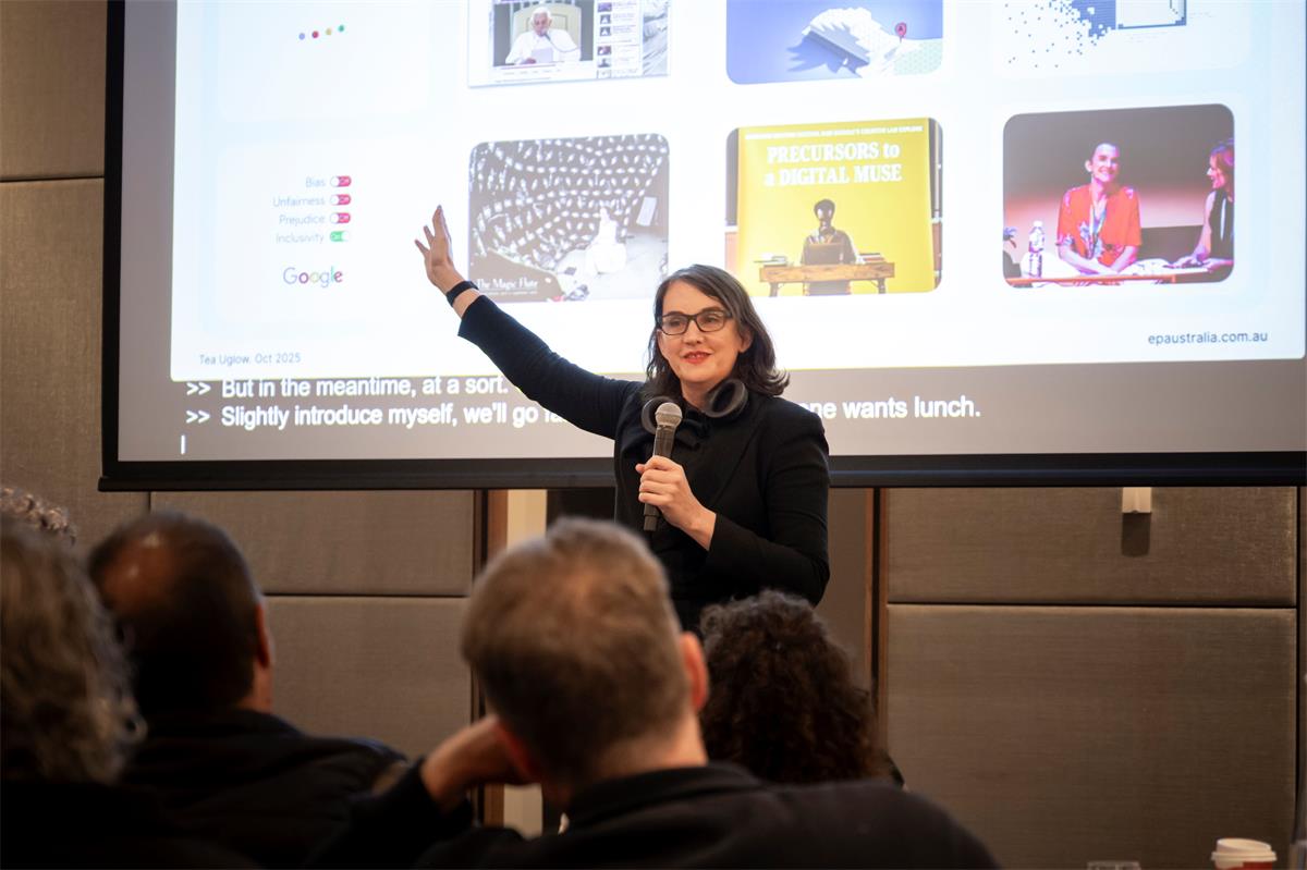 Tea Uglow, a woman dressed in black, holding a microphone and gesturing to a screen behind her