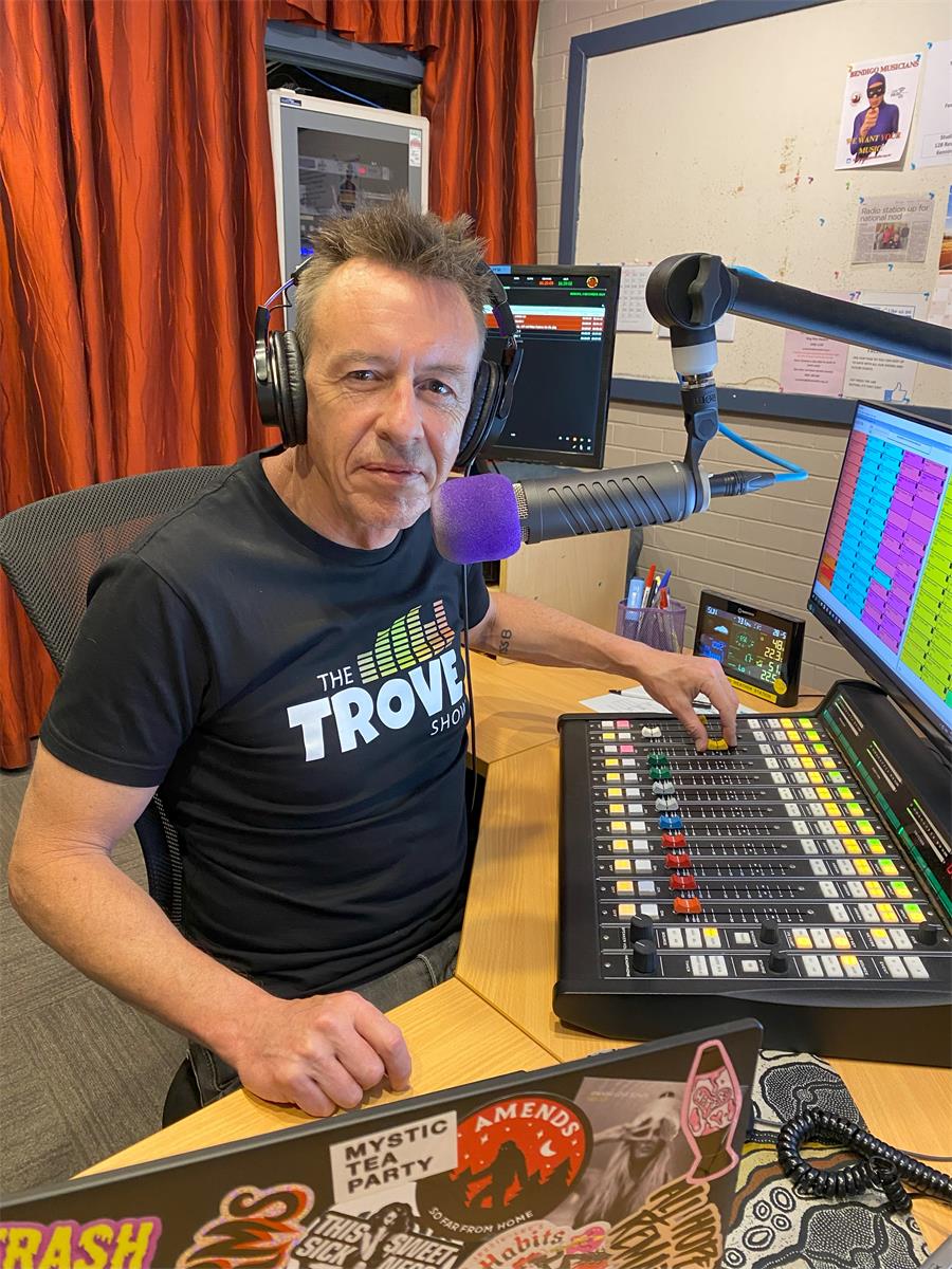 A person with headphones sits at a radio station, operating a soundboard with colorful buttons and a computer monitor in the background.