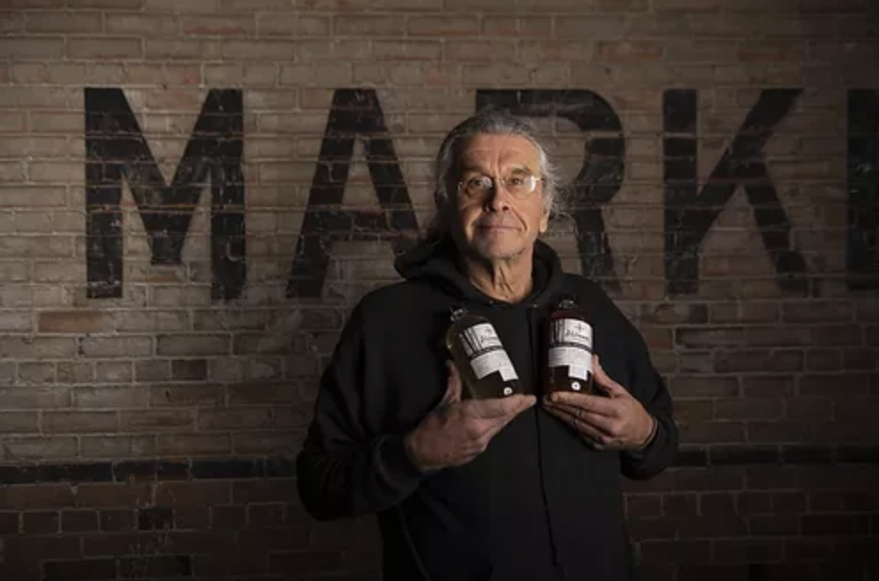 Paul Poutanen holding two bottles of his Alchemist Vinegar in front of a brick wall.