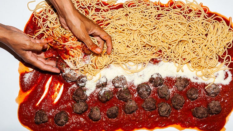 Components of spaghetti across a table being mixed by hands.
