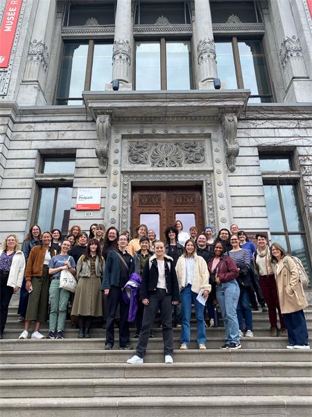 A group of smiling people wearing warm clothing stand on large stone steps that lead to an old museum with a stone facade and wooden doors.
