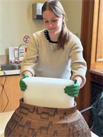A woman wearing nitrile gloves and a sweater holds a wide rectangular piece of foam that is partially in the opening of a very large basket. The basket is decorated with animals and patterns.
