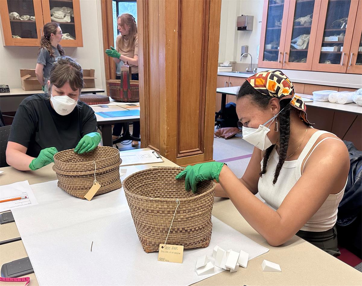 Two people wearing N-95 masks and nitrile gloves sit at a large work table working on baskets, with a stack of white wedges next to each basket.
