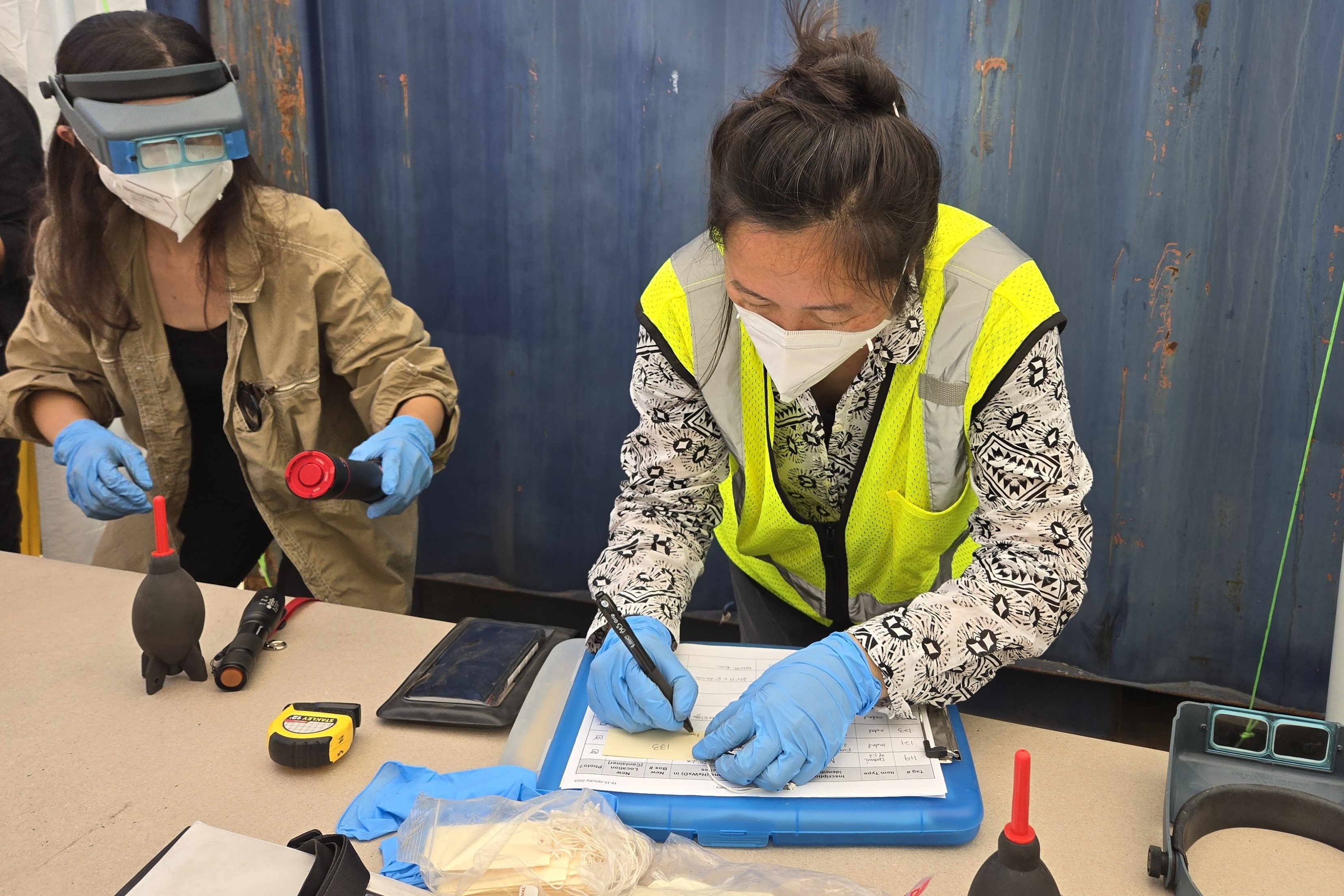 Two women in masks and gloves recording information on a clipboard