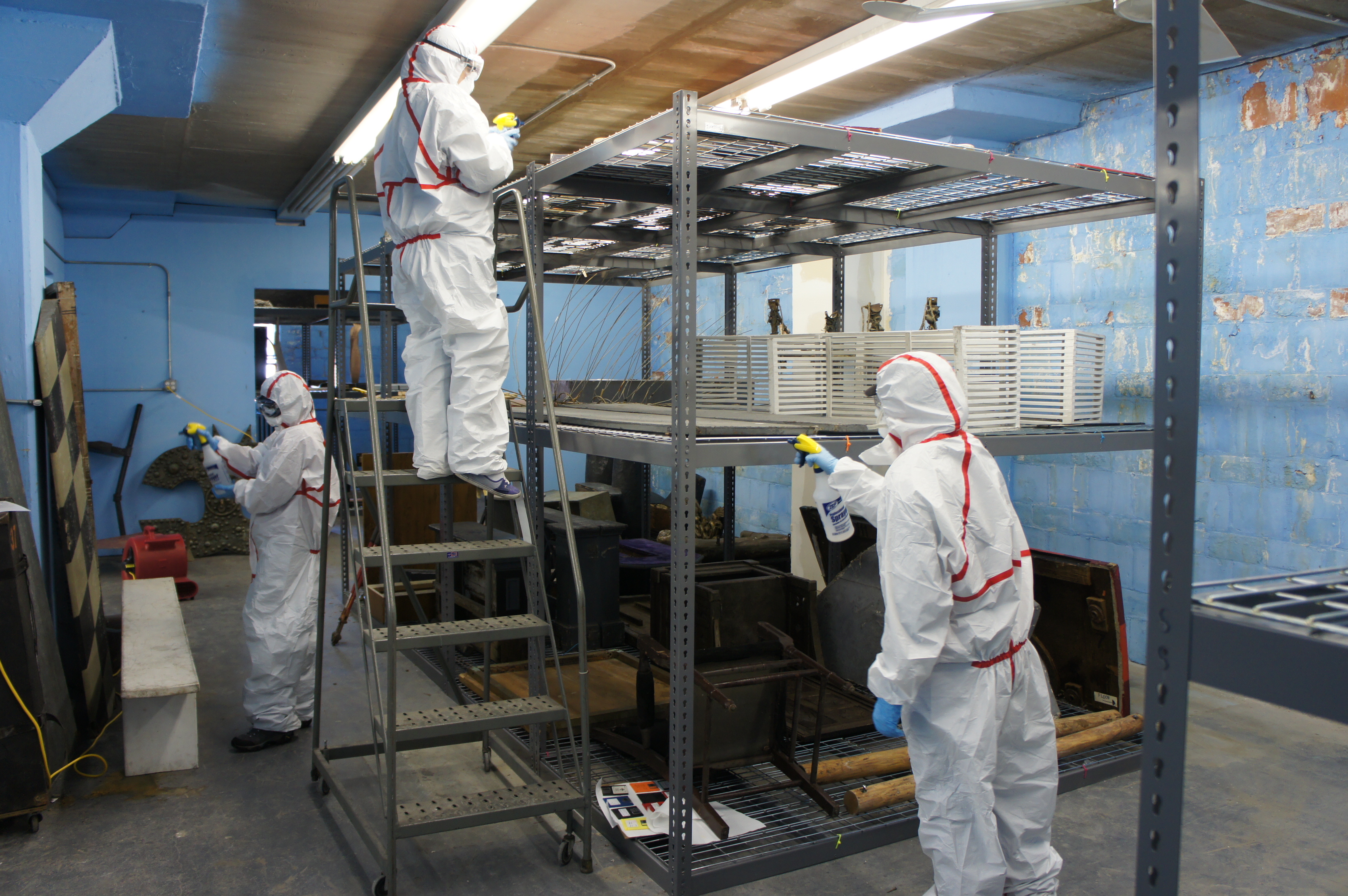Three people in Tyvek suits each holding a spray bottle in a space with shelving