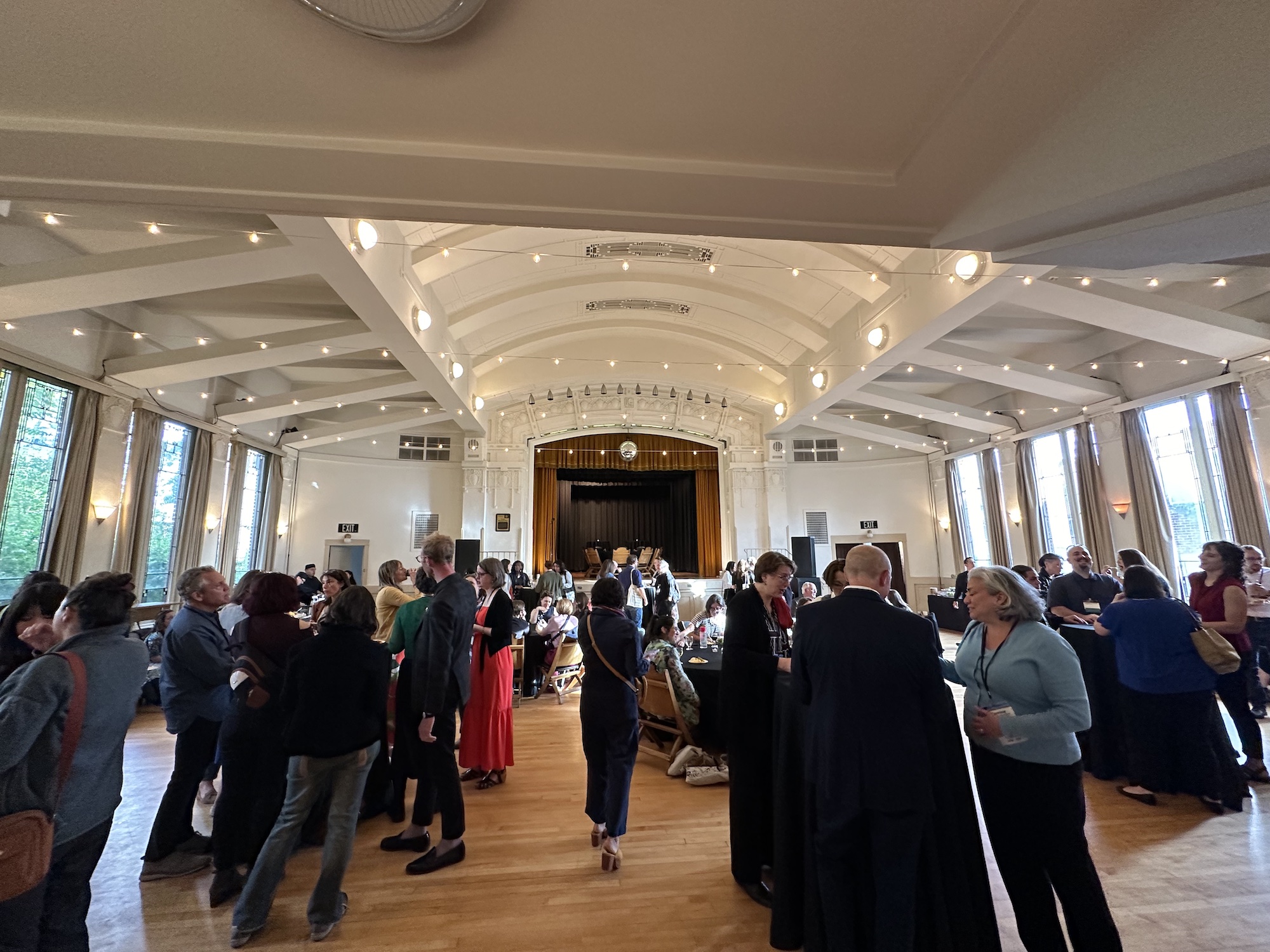 A large group of people stand inside a reception room with high ceilings, the people are eating, drinking, and talking.