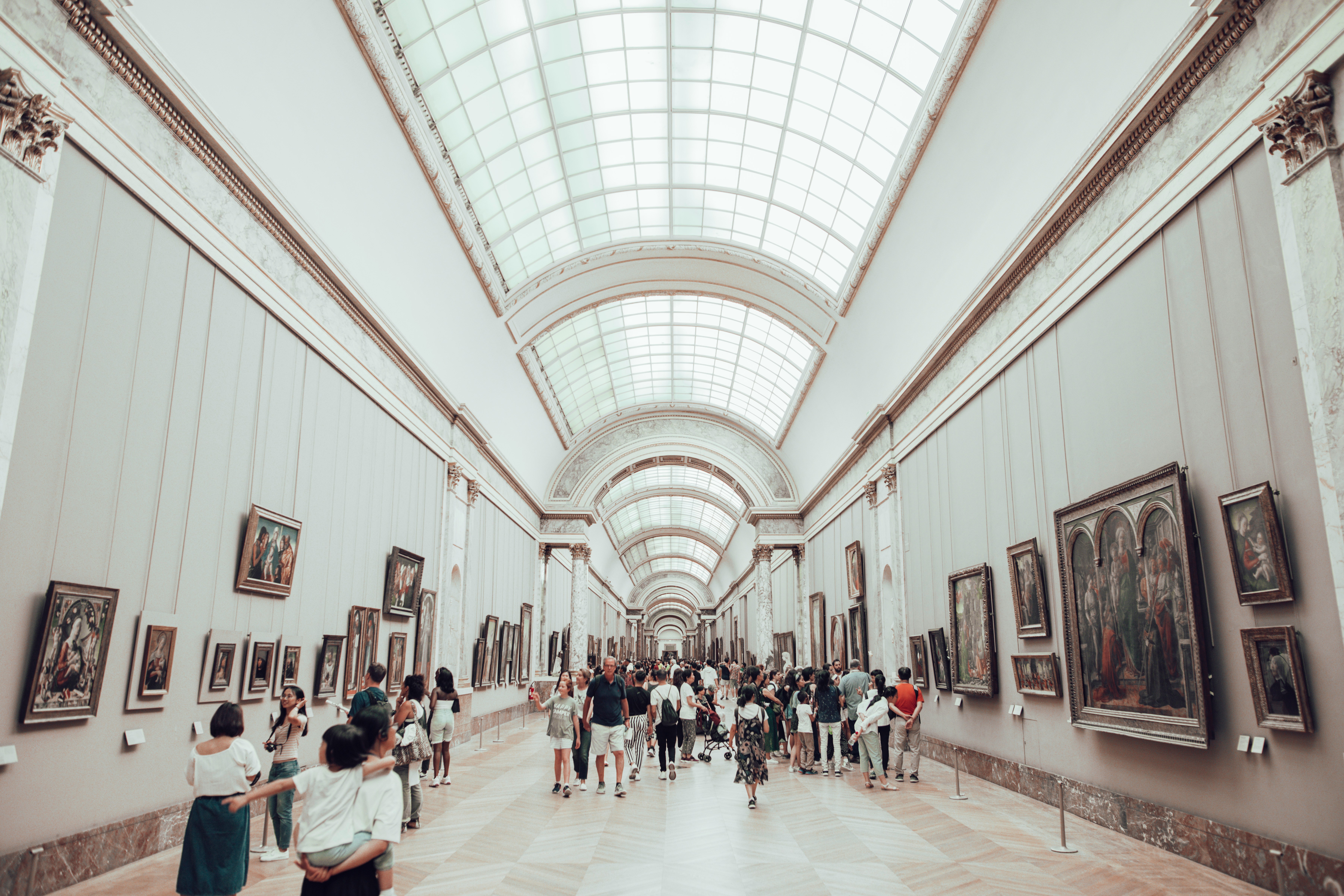 Patrons inside a museum gallery with high arched ceilings and rows of paintings on the walls