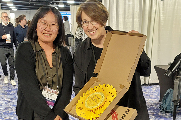 A woman smiles as a woman presents a paper sculpture in the shape of a Swiss cheese wheel as a poster award prize