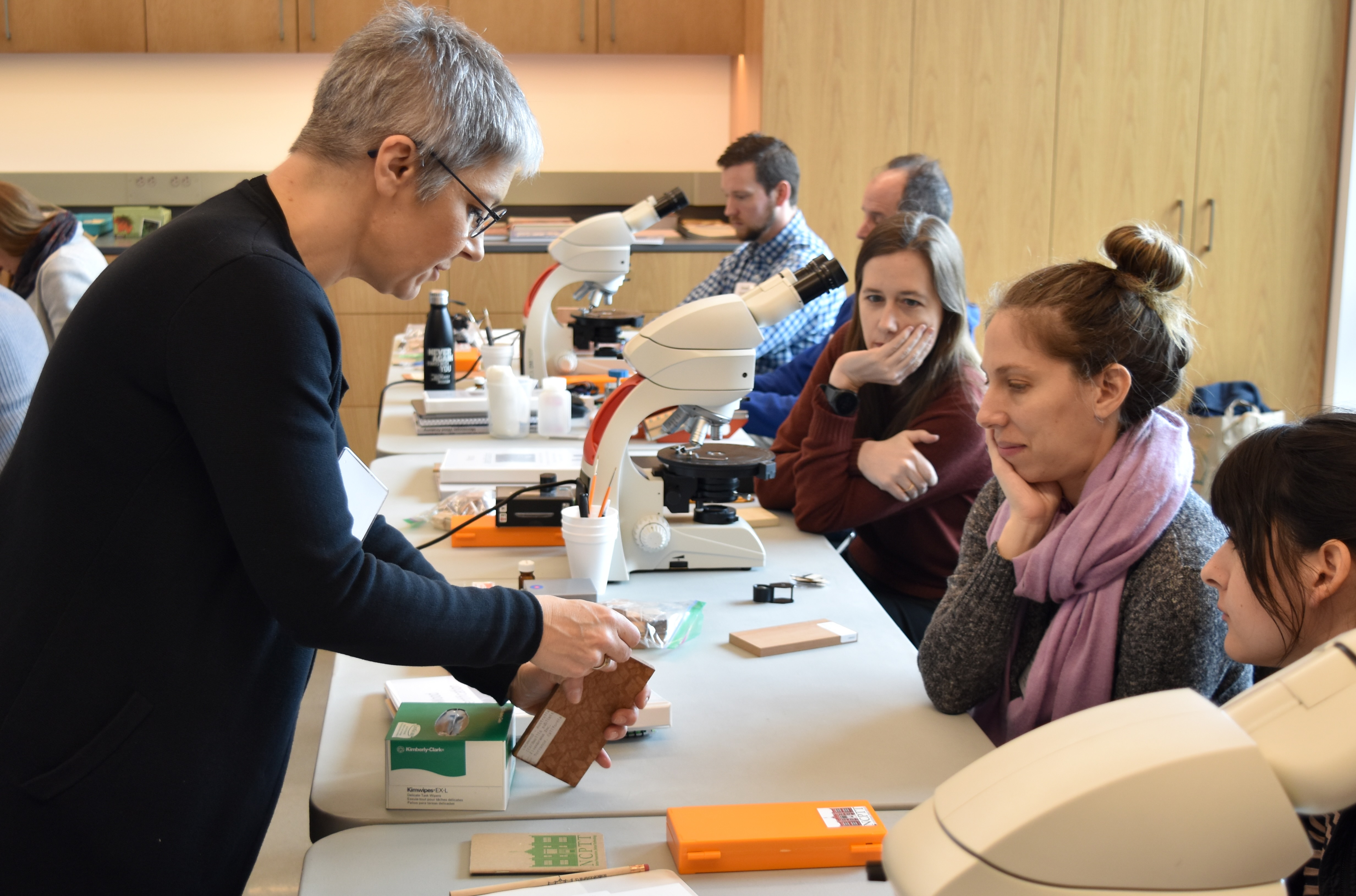 An instructor with short hair and glasses demonstrates a technique in front of a row of people in a workshop setting, with microshops in front of them