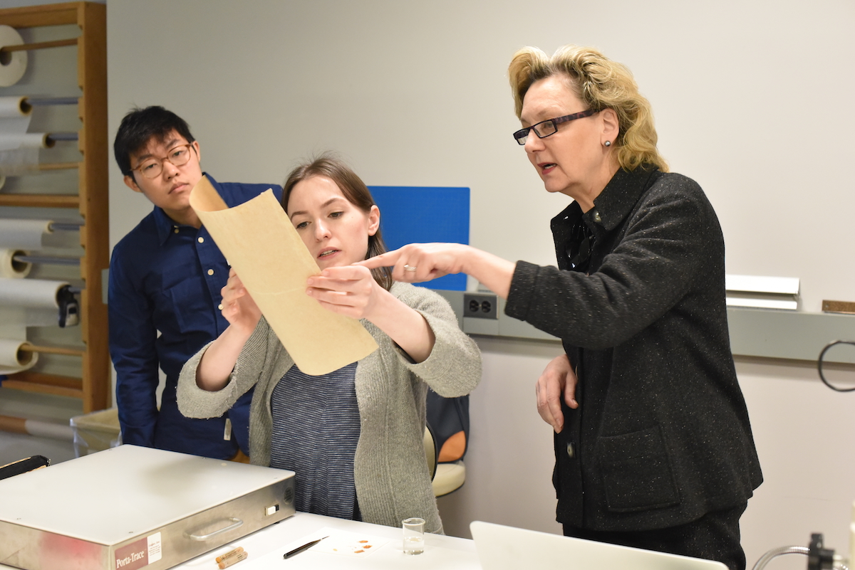 Instructor Peggy Ellis watches as students hold up a thick, yellowed paper for examination