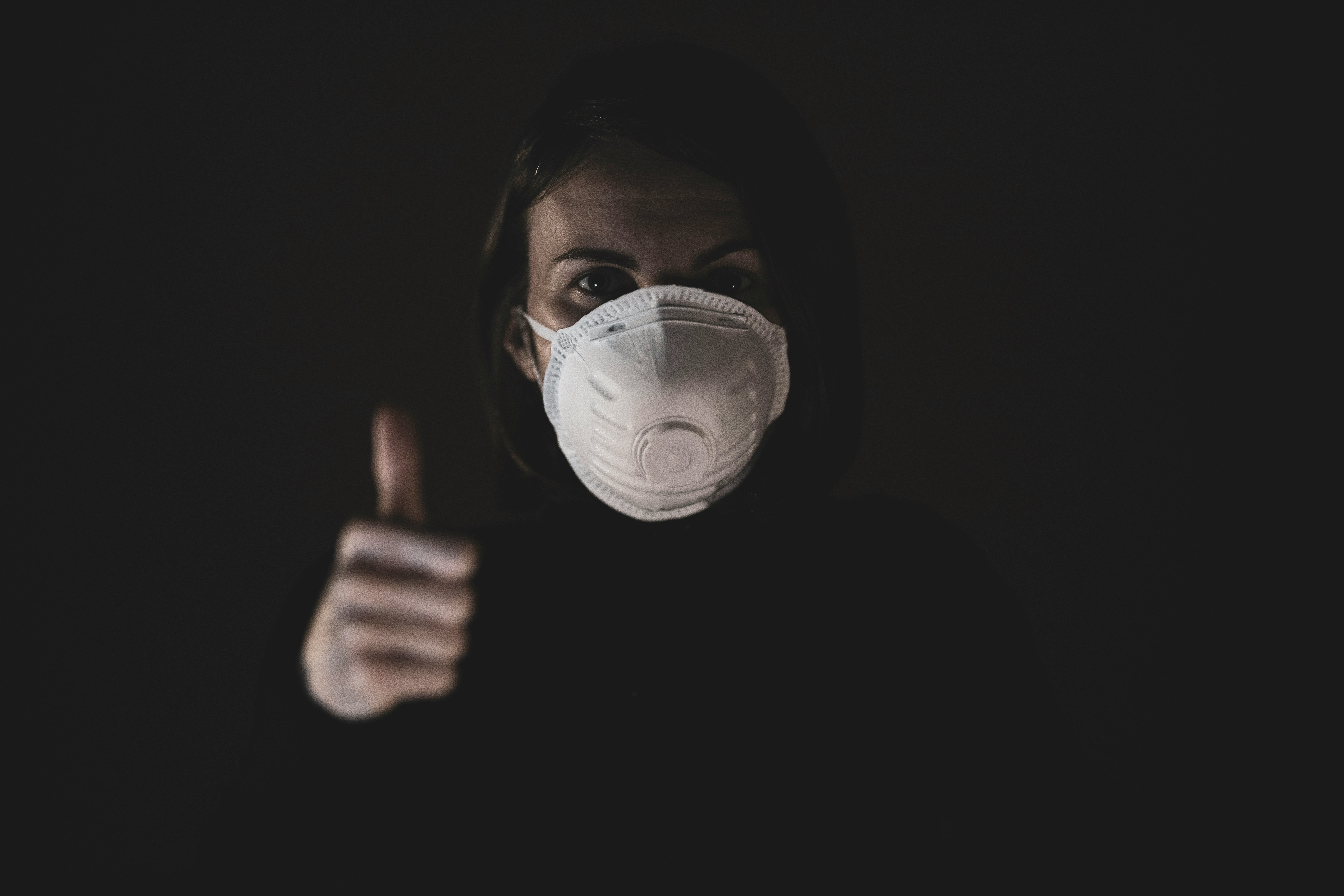 Dark photo of woman wearing a mask and showing thumbs up.